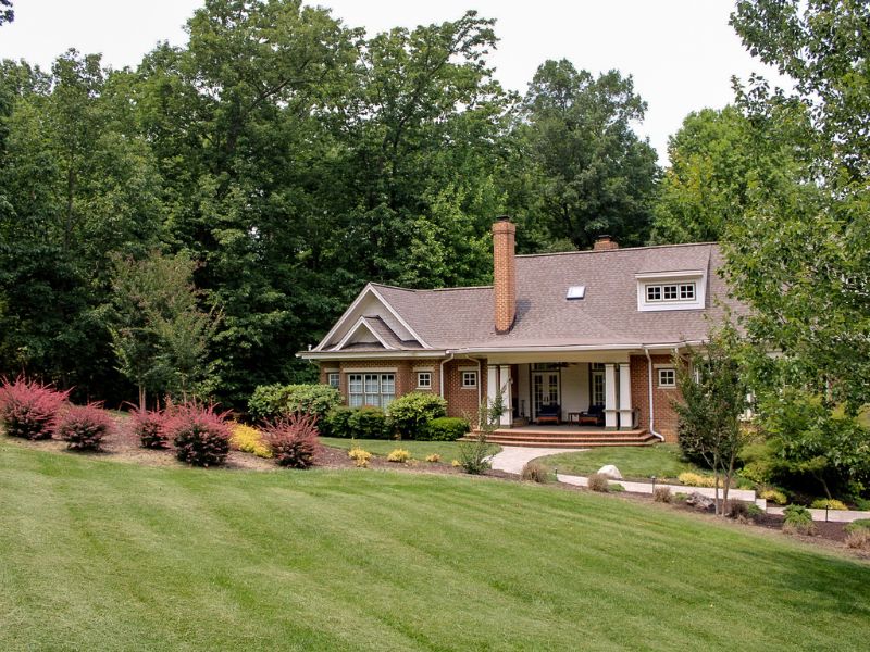 grounds-management-full-service-lawn-care-grass-homeowner Well-maintained front yard of a brick house surrounded by lush green trees, featuring freshly mowed grass, manicured shrubs, colorful plants, and clean landscaping, showcasing full-service lawn care.